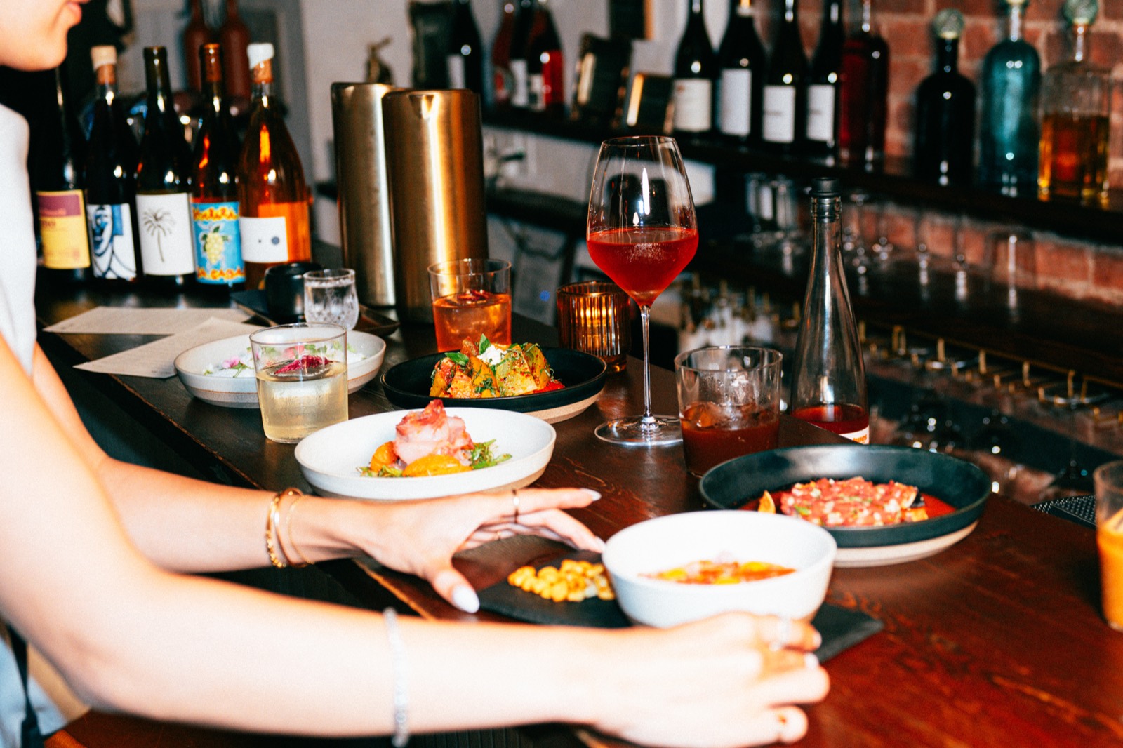 Share plates passed across the bar at WEST10WEST with natural wine bottles and a glass of sparkling rosé
