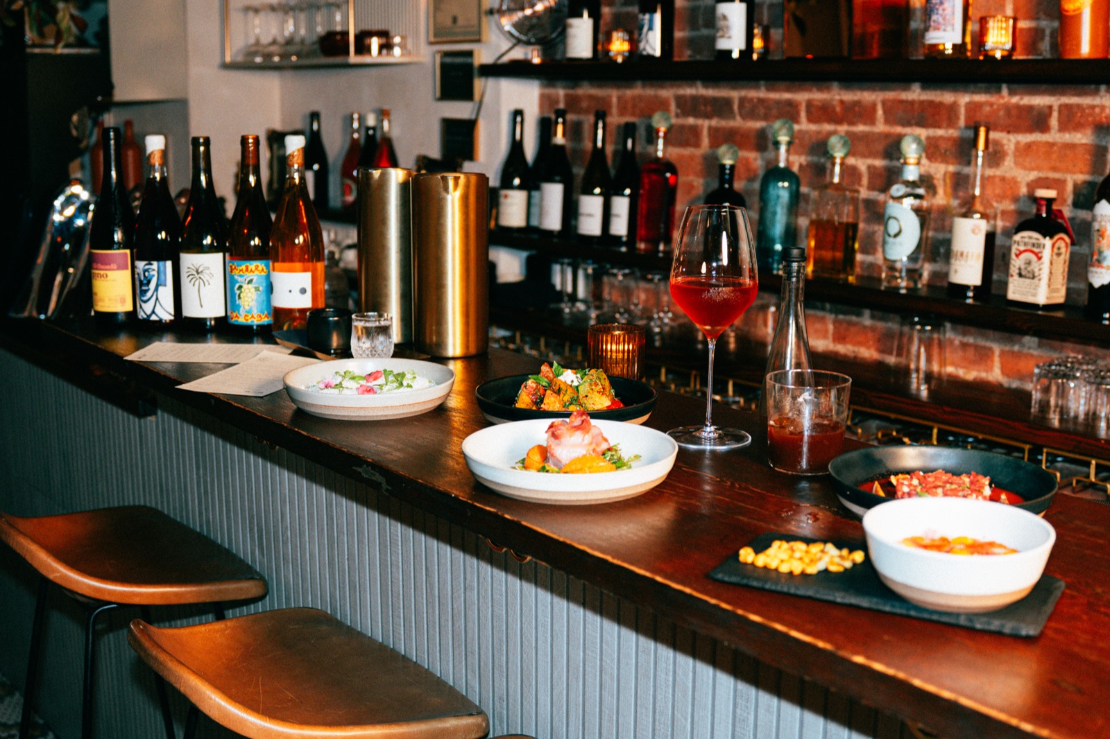 Share plates lined along the wood bar at WEST10WEST with shelves of natural wine bottles behind