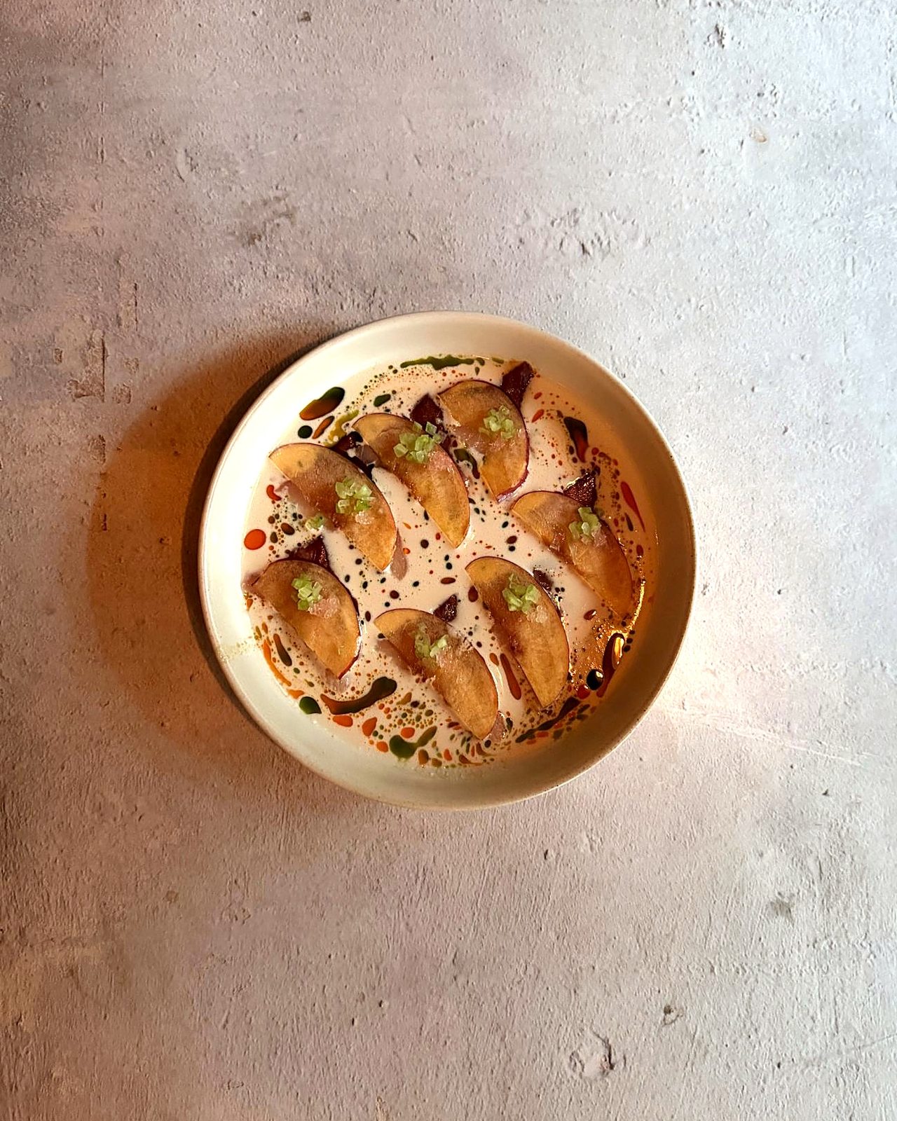 Overhead view of hamachi crudo with nectarine slices in ajo blanco, dotted with basil oil and chili oil, on a concrete table at WEST10WEST, a West Village share-plates restaurant