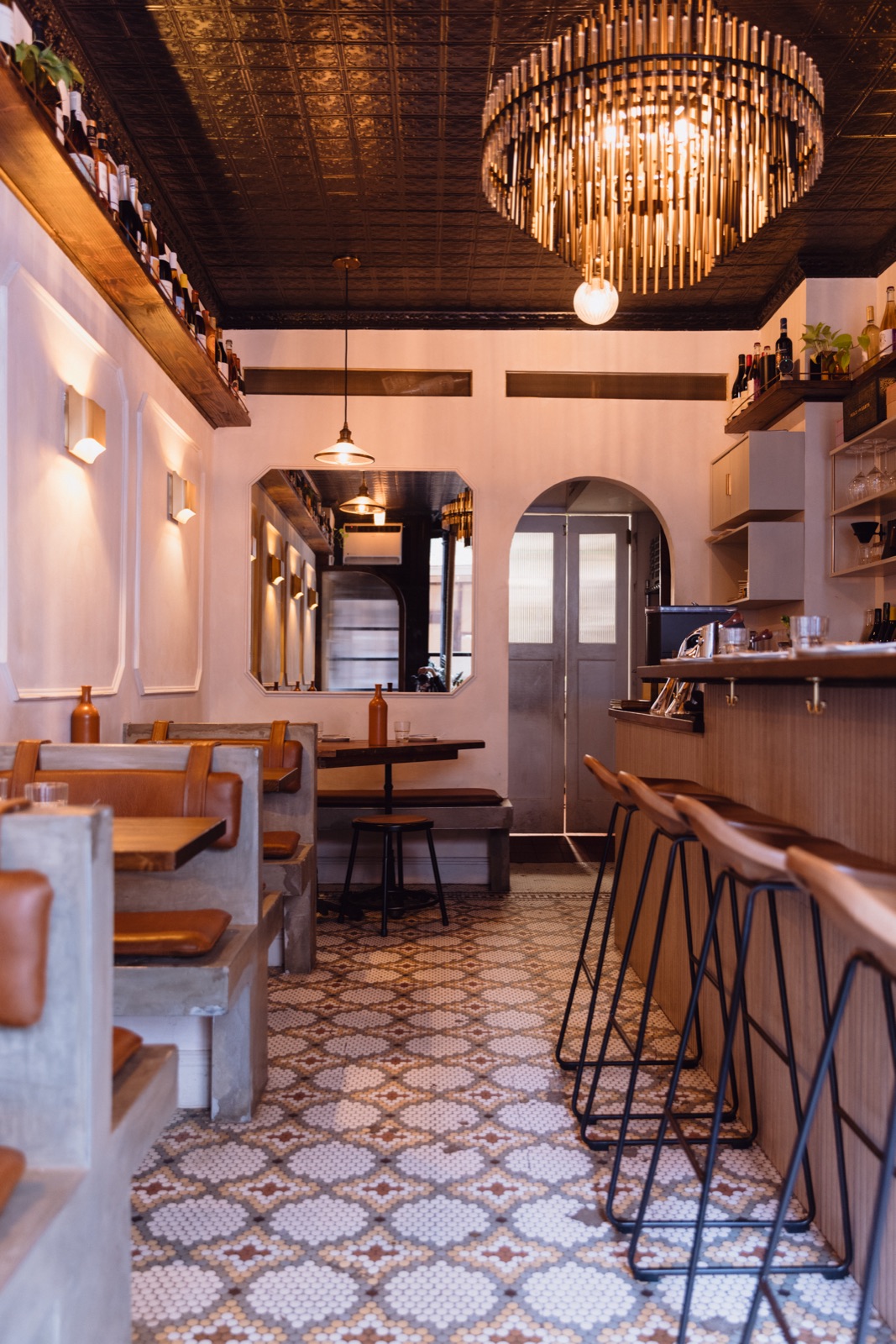 The WEST10WEST dining room in daylight: a tiered brass chandelier under a pressed-tin ceiling, mosaic tile floor, leather banquettes on the left and wood bar stools along the bar on the right