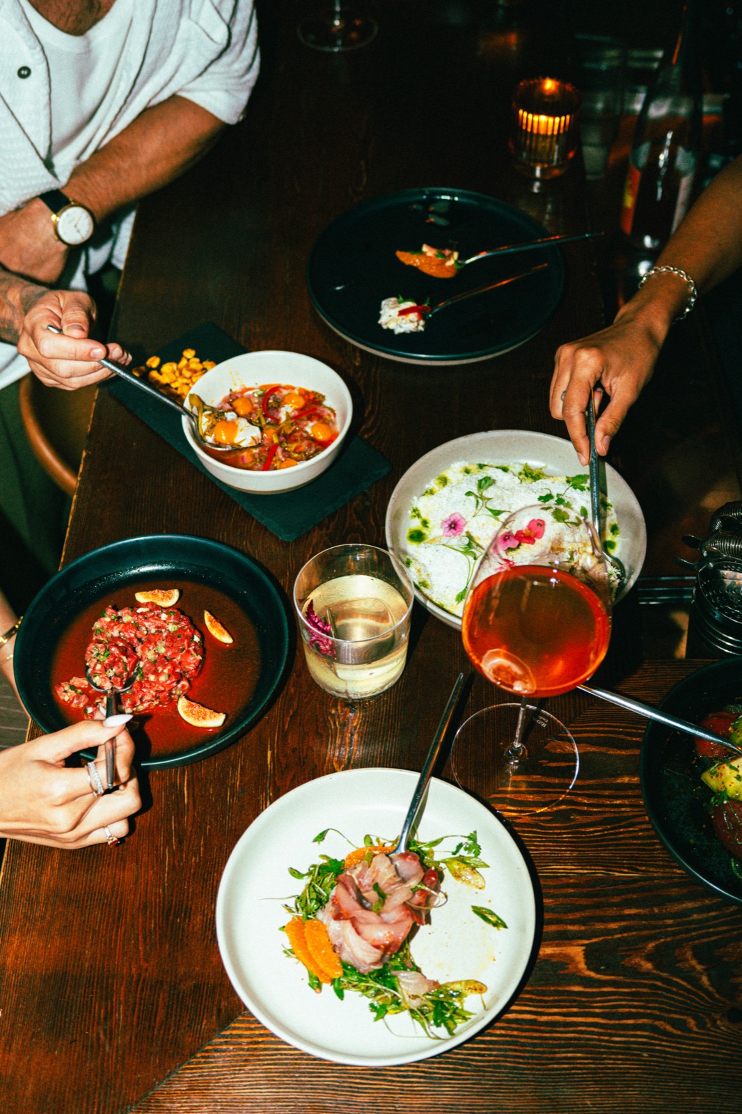 A wood table covered in share plates with sparkling rosé and cocktails, mid-dinner at WEST10WEST in the West Village