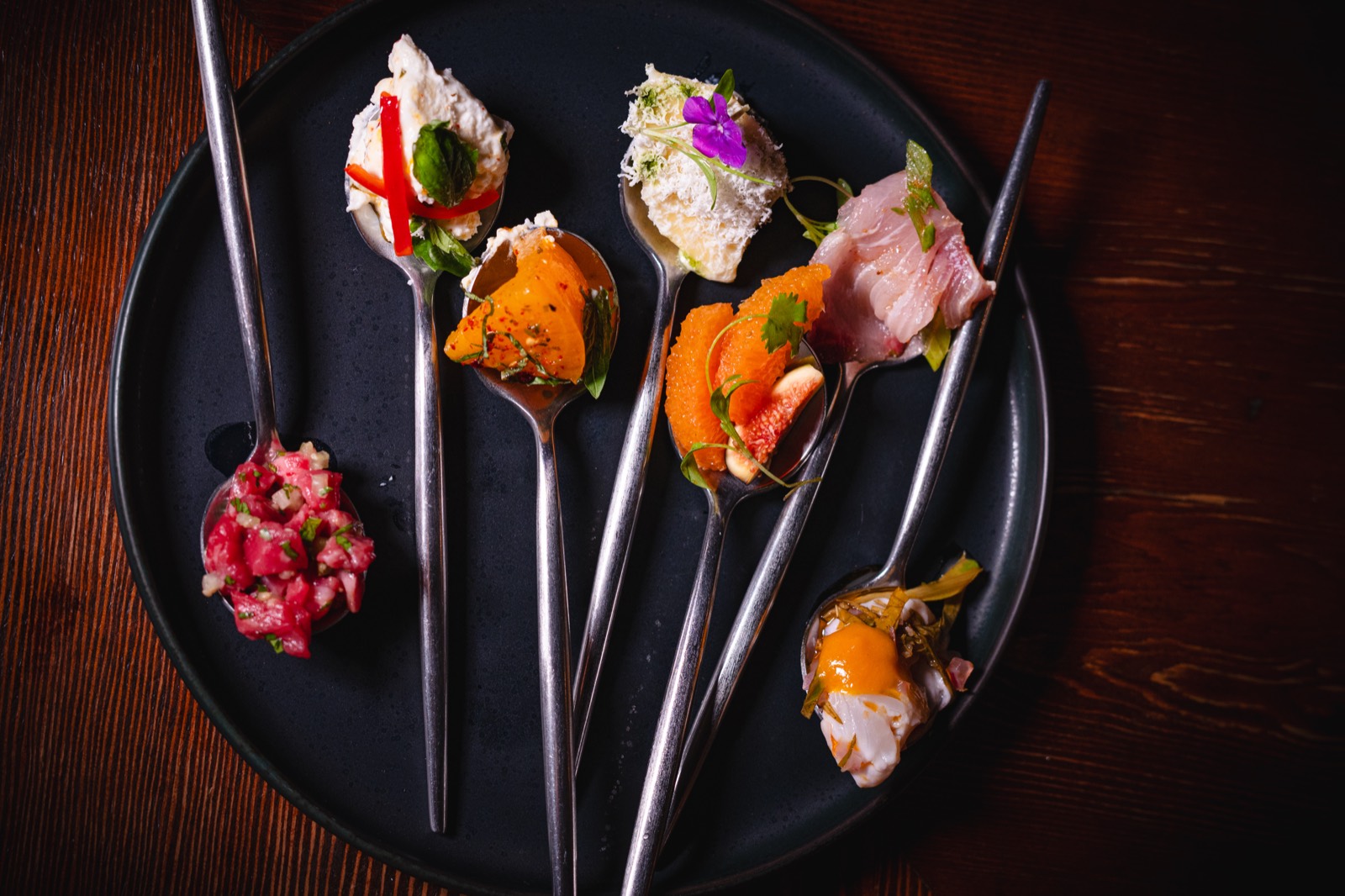 A flight of tasting spoons on a black plate at WEST10WEST — one bite of each share plate, from wagyu to crudo